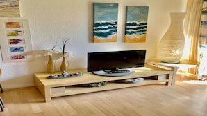 Een moderne woonkamer met een flatscreen televisie op een houten standaard, twee strandgerelateerde schilderijen aan de muur en een vaas met witte bloemen op een bijzettafeltje.