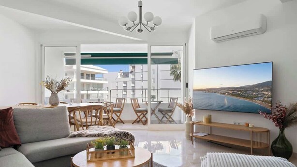 Ein modernes Wohnzimmer mit Sofa, Couchtisch und Fernseher. Ein Balkon bietet Blick auf das Meer.