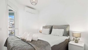 Ein ordentlich gemachtes Bett mit einer gesteppten Bettdecke, einer Nachttischlampe und Blick durch ein offenes Fenster hinaus.
