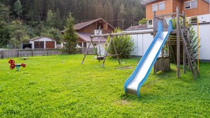 Un parque infantil con un tobogán y columpios, rodeado de césped y una casa con terraza de madera.