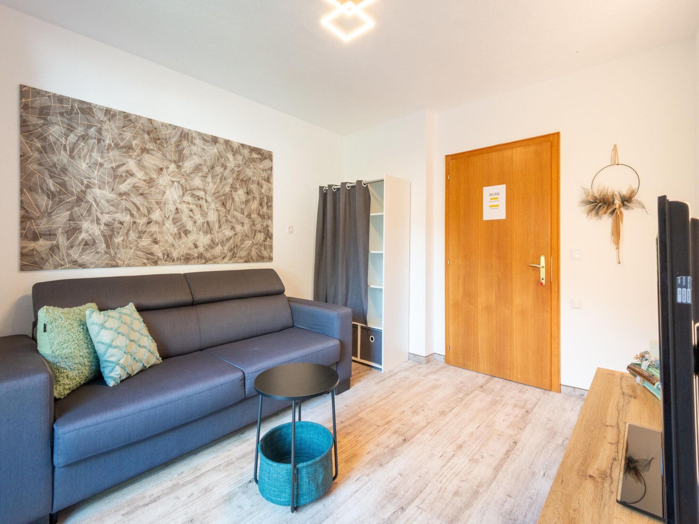 Ein modernes Wohnzimmer mit einem grauen Sofa, einem kleinen runden Tisch, einem blauen gewebten Korb und einer Holztür mit einem Schild.