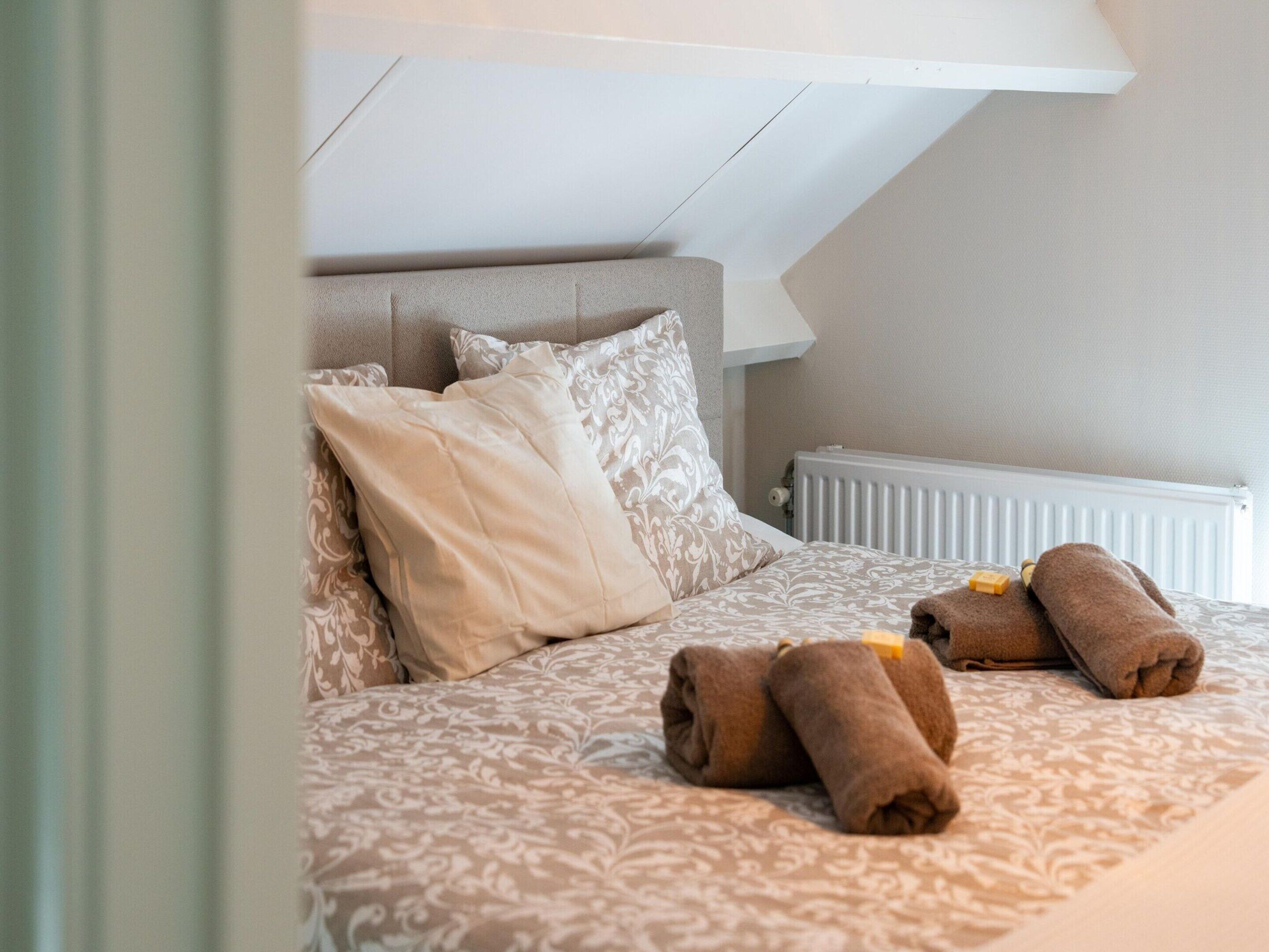 A neatly made bed with patterned bedding, a headboard, and rolled towels on the bed.