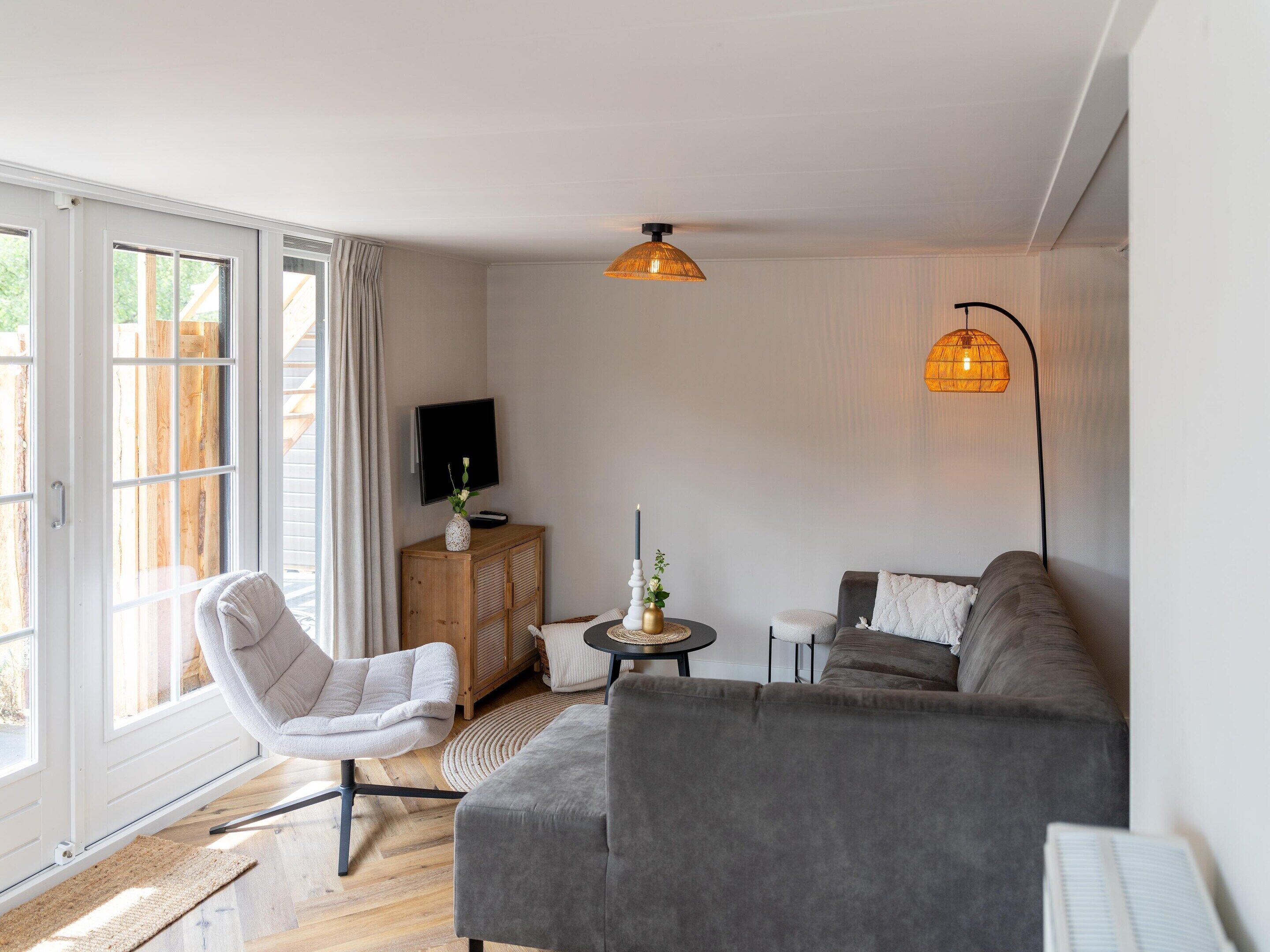 Ein Wohnzimmer mit grauem Sofa, einem weißen Sessel, einem Fernseher auf einer Holzkommode und einer Stehlampe.