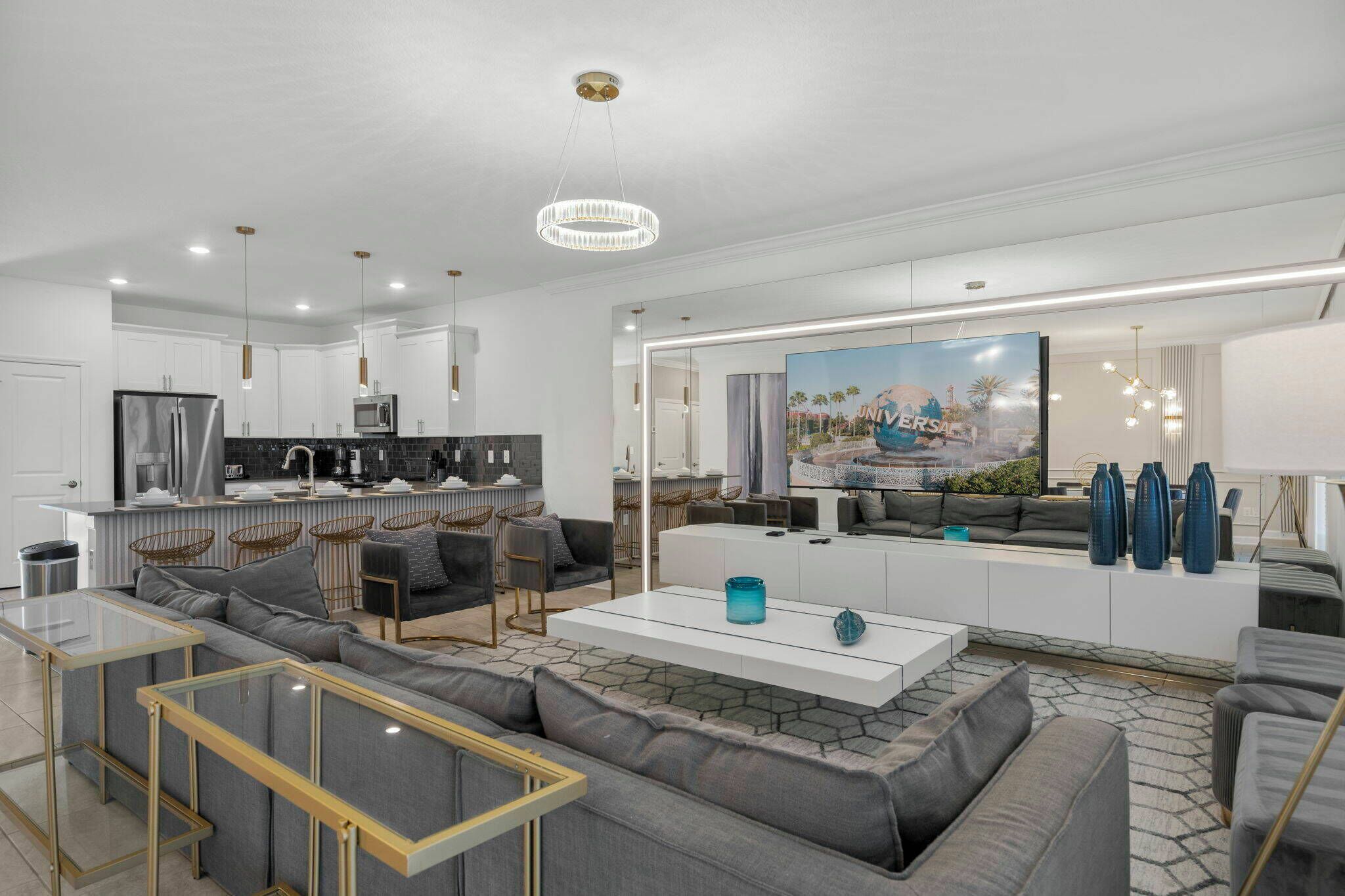 A modern living room with a large sectional sofa, a glass coffee table, and a kitchen area with white cabinets and a large island.