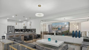 Una sala de estar moderna con un sofá seccional grande, una mesa de centro de vidrio y una cocina con gabinetes blancos y una isla grande.