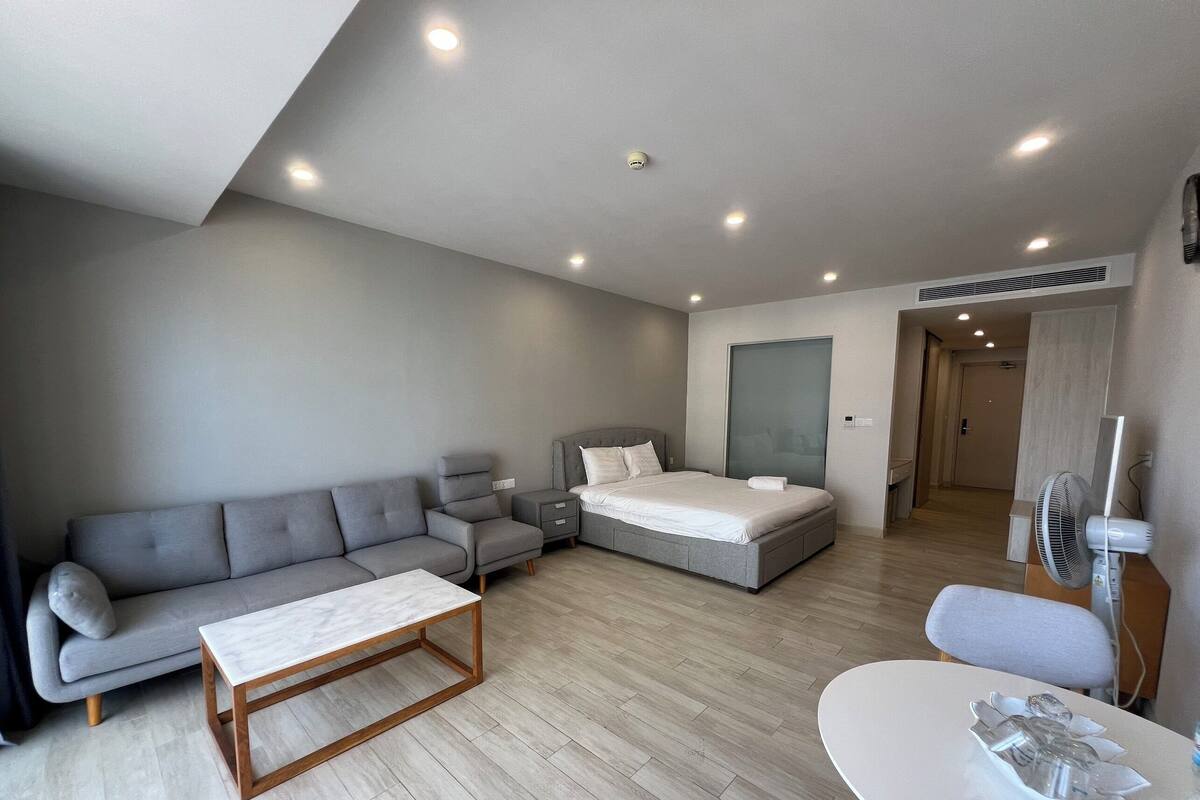 A modern, well-lit bedroom with a grey sofa, a bed, a small bedside table, and a white table with a decorative item.