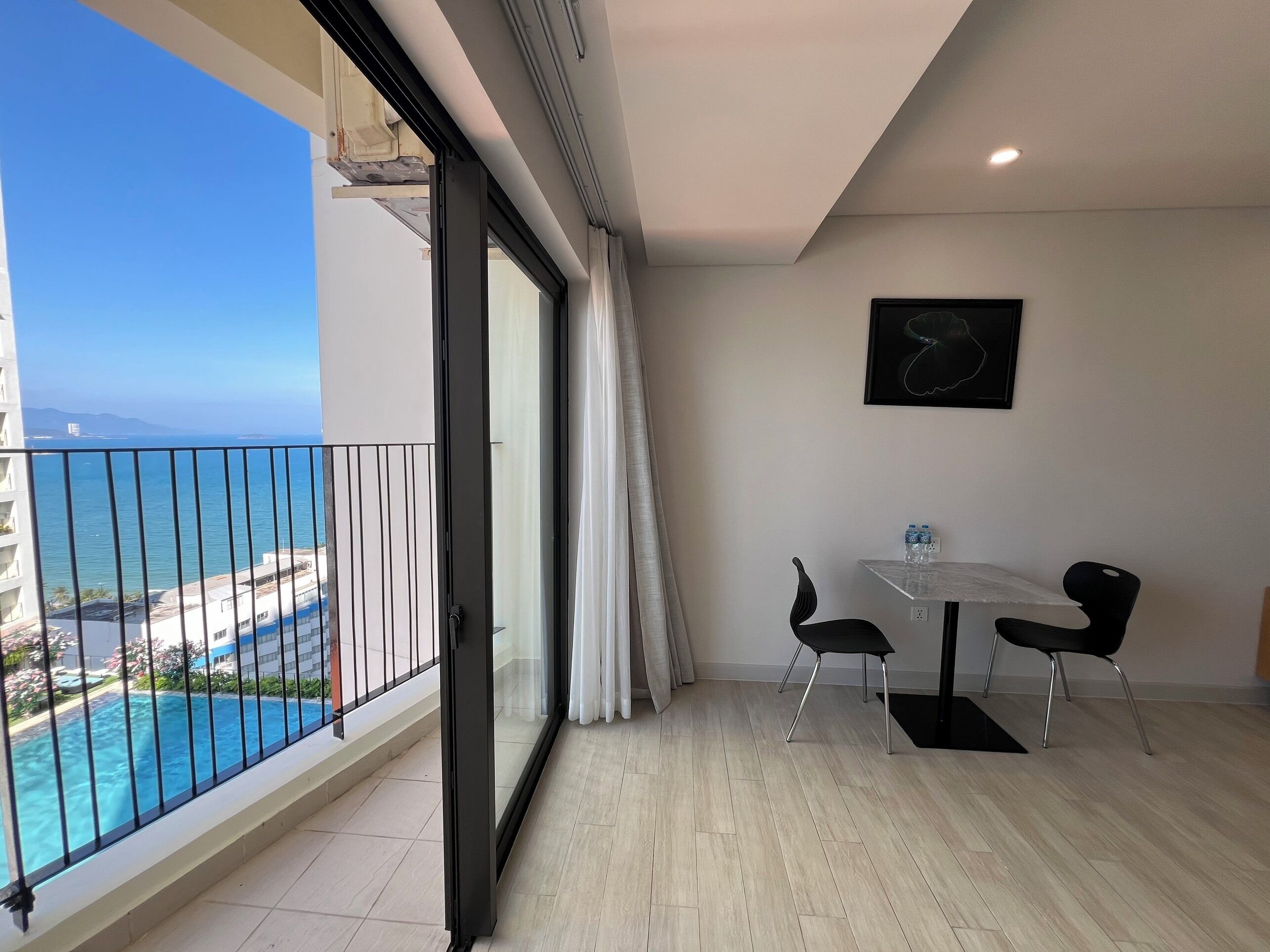 Un balcón con vistas al mar y a una piscina. Hay una mesa con sillas y una botella de agua encima.