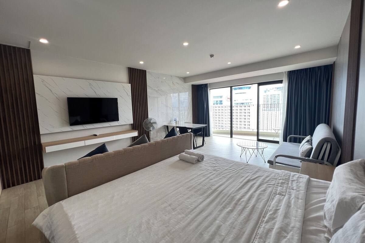 A modern hotel room with a large bed, a flat-screen TV mounted on the wall, a desk with a chair, and a balcony with a view of the city.