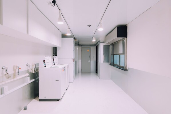 A clean, modern hallway with white walls and floors, featuring a laundry area with industrial-style machines, a window on the right, and a door at the end. - Minimalist Loft 405 (Osaka)