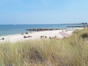 Une plage où des gens se prélassent au soleil et se baignent, une digue rocheuse et un voilier à l’horizon.