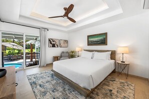 A bedroom with a bed, two bedside lamps, a ceiling fan, a painting, and a view of an outdoor pool area.
