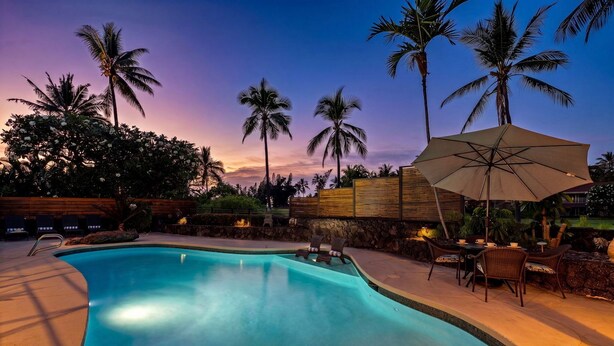 Une piscine entourée de palmiers, avec des chaises longues et un parasol, le tout baigné par la lumière d’un coucher de soleil.