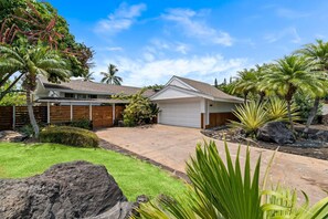 Una casa de dos pisos con garaje, rodeada de follaje tropical y grandes rocas.