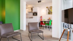 Una sala de estar moderna con dos sillones de cuero, piso de madera, una pared verde de acento, una zona de comedor con mesa y sillas, una cocina al fondo y un televisor sobre un atril.