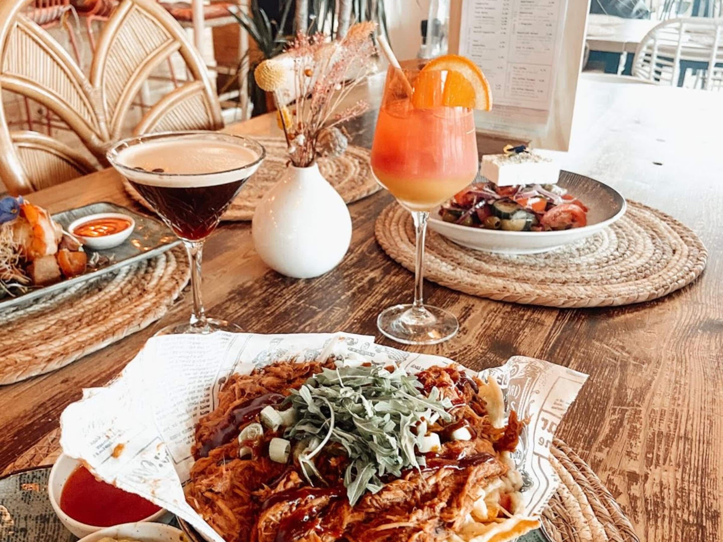 A wooden table with a variety of dishes, including a bowl of shredded meat with greens, a glass of red drink with an orange garnish, and a plate of vegetables.