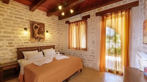 A bedroom with a stone wall, wooden ceiling beams, a bed with white linens, a painting on the wall, and a window with curtains. - Holiday Home in Arkadi With Private Pool (Rethymno)