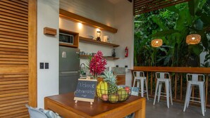 Une table en bois, un tableau noir, un panier de fruits et une plante en pot dans un espace intérieur moderne comprenant un coin cuisine et offrant une vue sur la verdure.