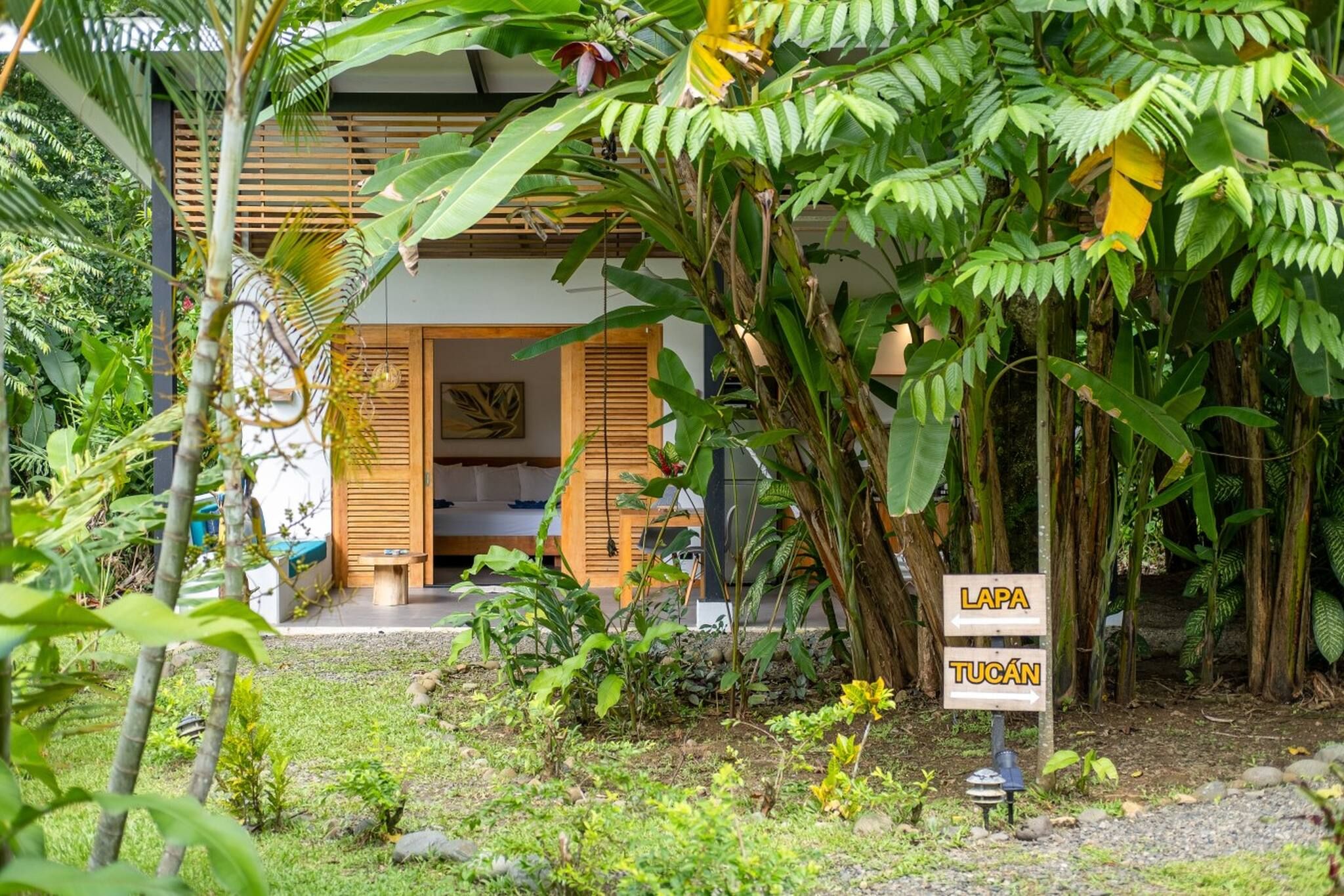 Un jardin tropical avec une maison en bois, un lit et un panneau indiquant les directions vers Lapa et Tucan.