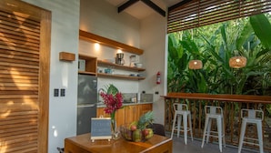 Une cuisine moderne dotée d’un réfrigérateur encastré, d’étagères en bois et d’une machine à café. On y trouve également un coin repas avec une table et des chaises, et une vue sur une végétation luxuriante à l’extérieur.