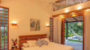 A bedroom with a bed, a nightstand, a wall-mounted lamp, a ceiling fan, and a view of a garden with a hammock.