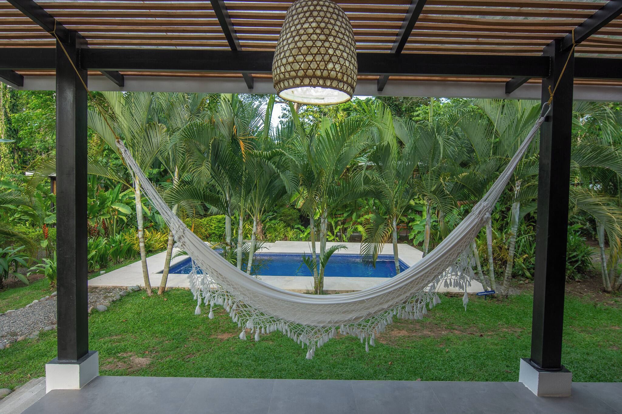 A hammock under a wooden pergola with a pool in the background surrounded by lush greenery.