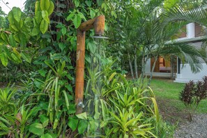 A wooden water feature with a spout, surrounded by lush greenery and a white building in the background.
