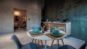 Un coin repas avec une table ronde, des chaises bleues et de la vaisselle vert turquoise. On aperçoit en arrière-plan une cuisine équipée d’un évier et de placards.