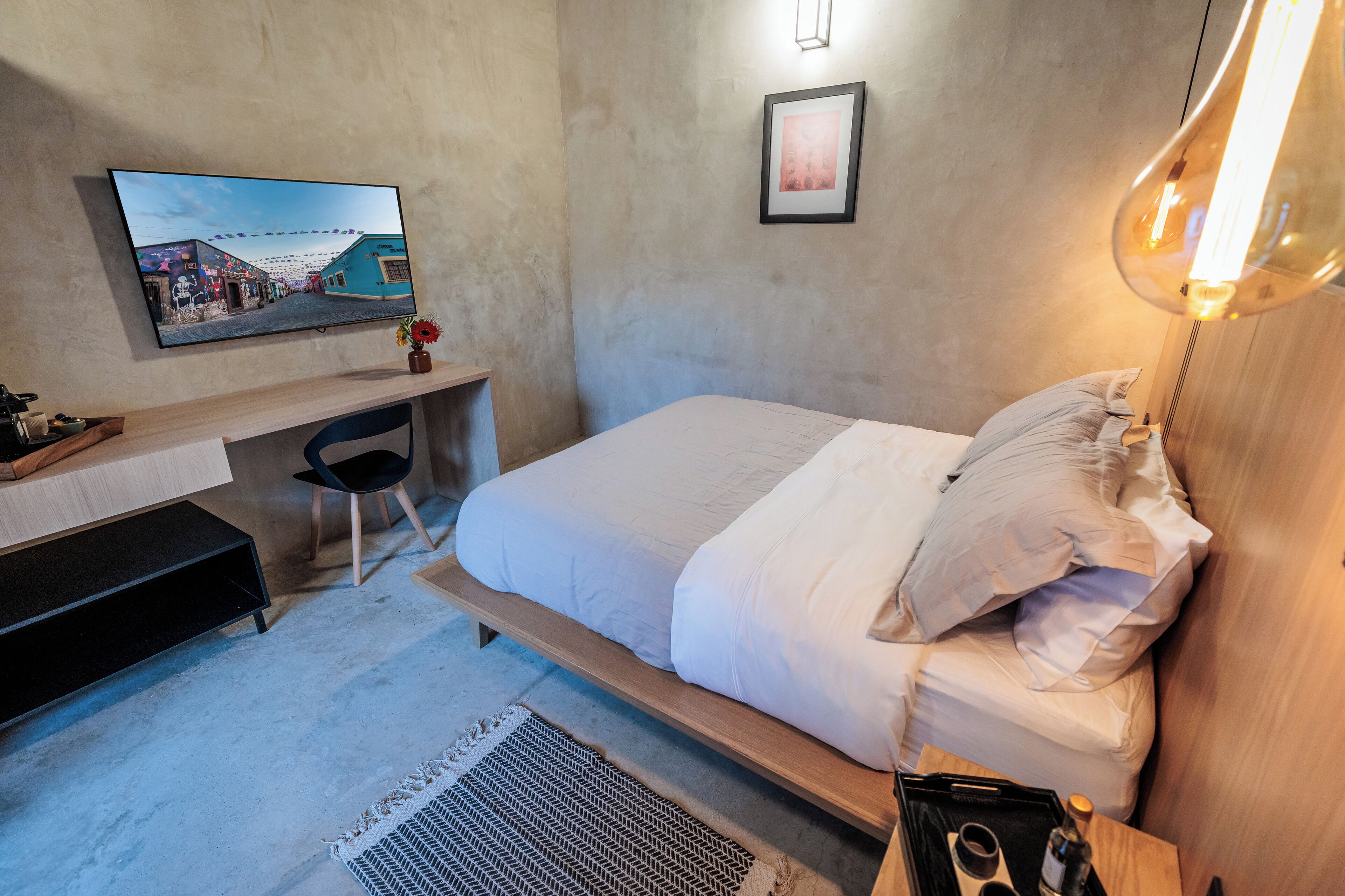 A modern bedroom with a bed, bedside table, lamp, TV, desk, chair, and a small plant.