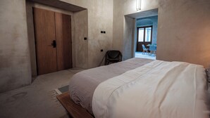 Une chambre avec un grand lit, une armoire en bois et une chaise. Il y a une fenêtre et une porte qui donne sur une autre pièce.