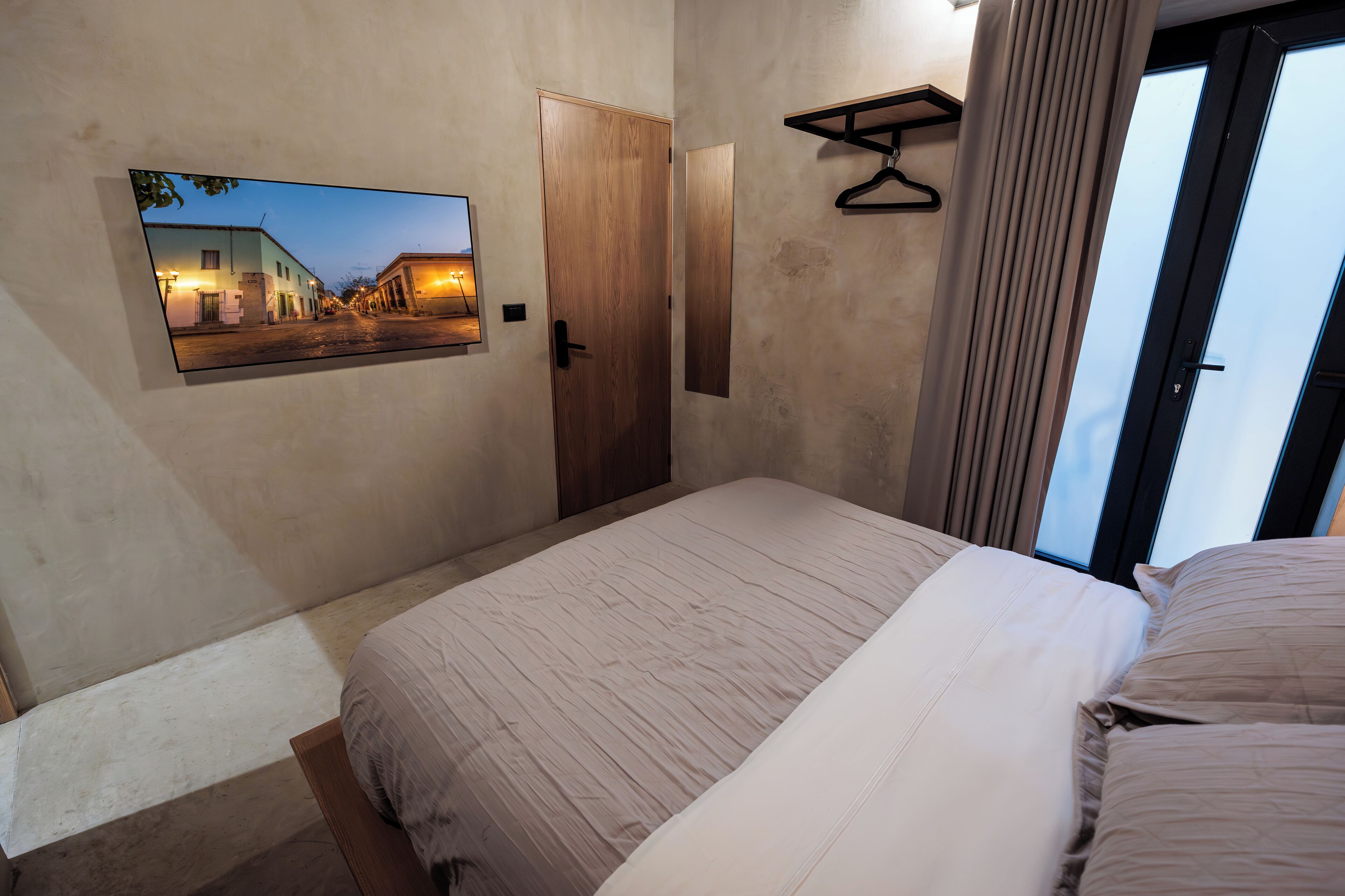 Ein Schlafzimmer mit einem Bett, einer Tür, einem Kleiderständer und einem an der Wand befestigten Fernseher, der eine Straßenszene zeigt.