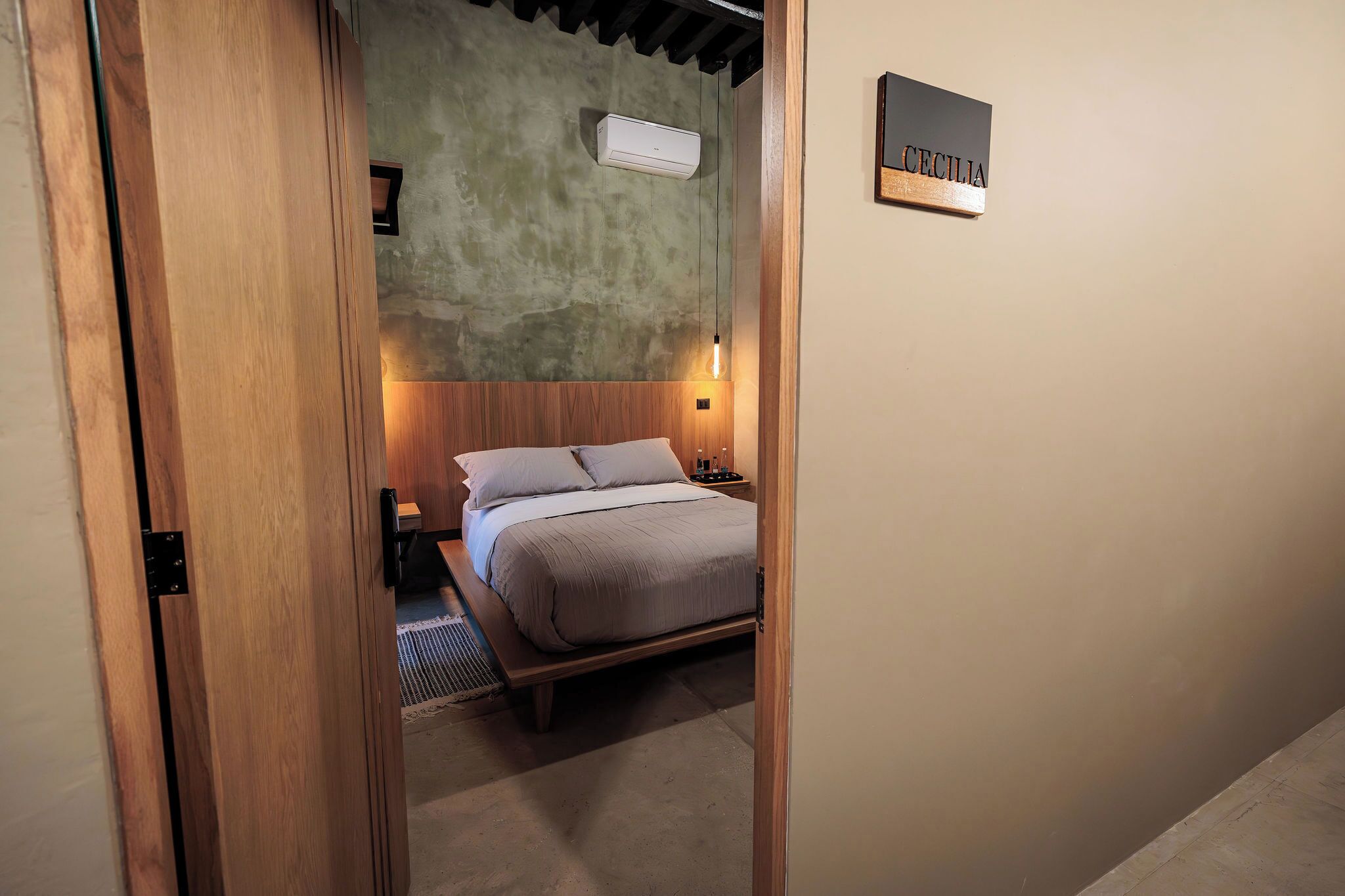 A bedroom with a wooden bed, a nightstand, a wall-mounted air conditioner, and a nameplate on the wall.