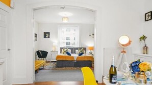 Ein modernes Hotelzimmer mit Holzboden, einem gelben Sessel, einem Bett mit orange- und blau gestreuter Bettwäsche, einem kleinen Tisch mit Flasche und Gläsern sowie einem Spiegel an der Wand.