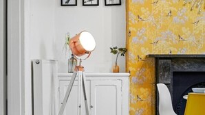 Ein Raum mit einer auf einem Stativ stehenden Lampe, einem weißen Schrank, floralem, gelbem Wandpapier, einem weißen Stuhl und einem Kamin.