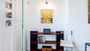 A hallway with a white door, a wall-mounted picture, a shelf with books, a vintage suitcase, and a hanging white cushion.
