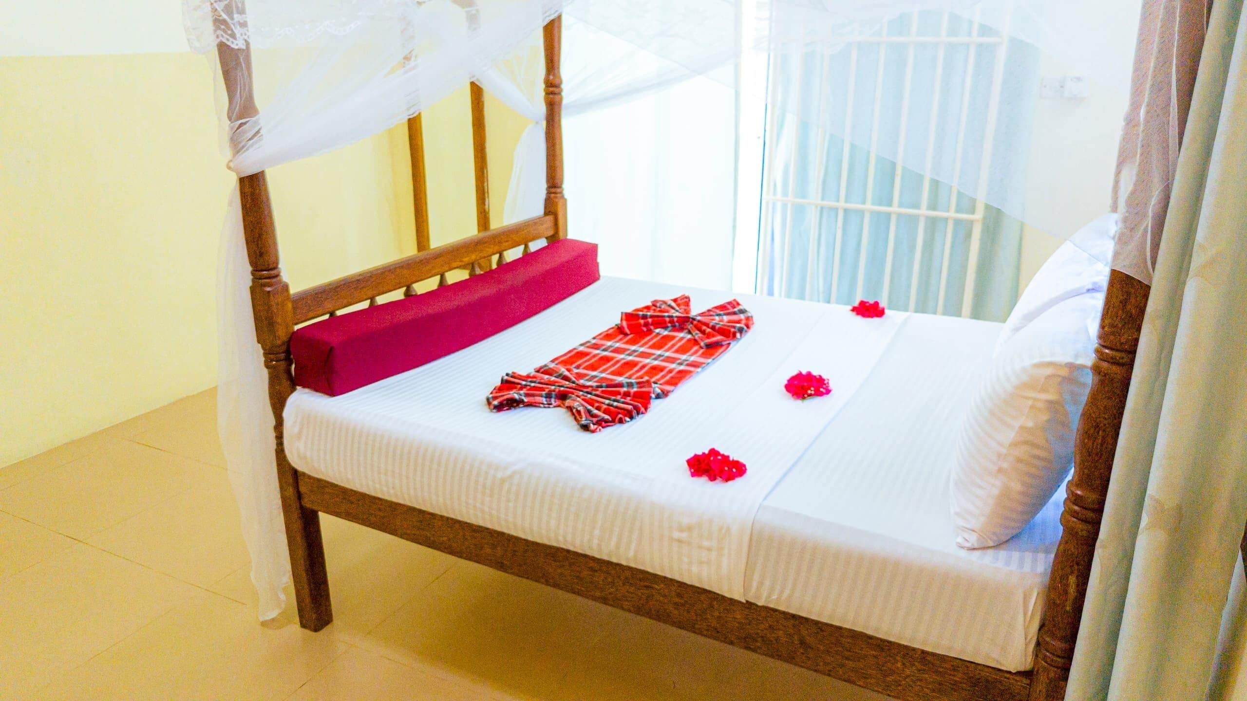 A bed with a wooden frame, white bedding, a red pillow, and a red and white checkered blanket.