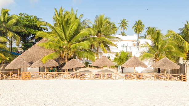 A beach with thatched umbrellas and palm trees, a white building in the background, and a sandy beach. - Sands inn Hotel (Pwani Mchangani)