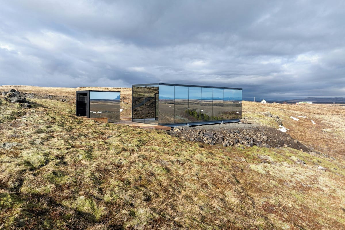 A modern building with a glass facade reflecting the landscape, situated on a grassy hill with a cloudy sky.