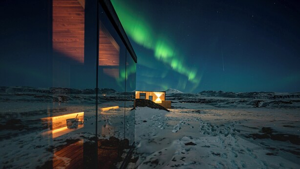 Schneebedeckte Landschaft mit einer Holzstruktur, die Licht reflektiert, bewölkter Himmel und Polarlichtern.