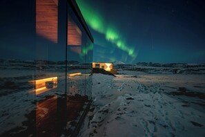 Un paysage enneigé, avec une structure en bois qui réfléchit la lumière, un ciel dégagé et des aurores boréales.
