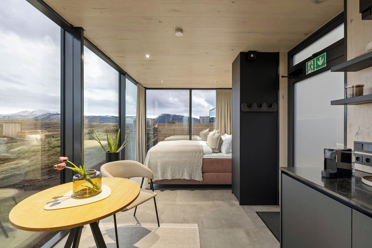 A modern hotel room with a large window, a bed, a small table with a vase, and a kitchenette.