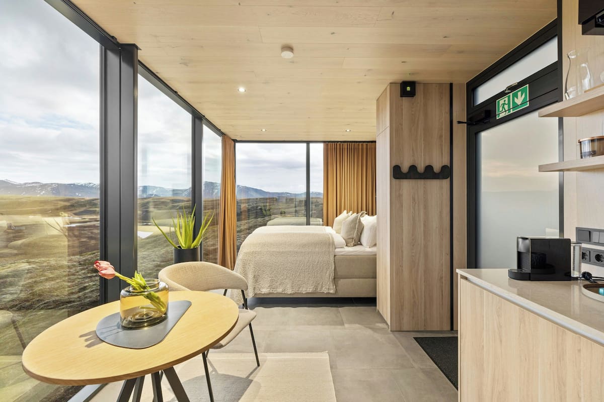 A modern room with a large window, a bed, a dining area with a table, and a kitchen area.