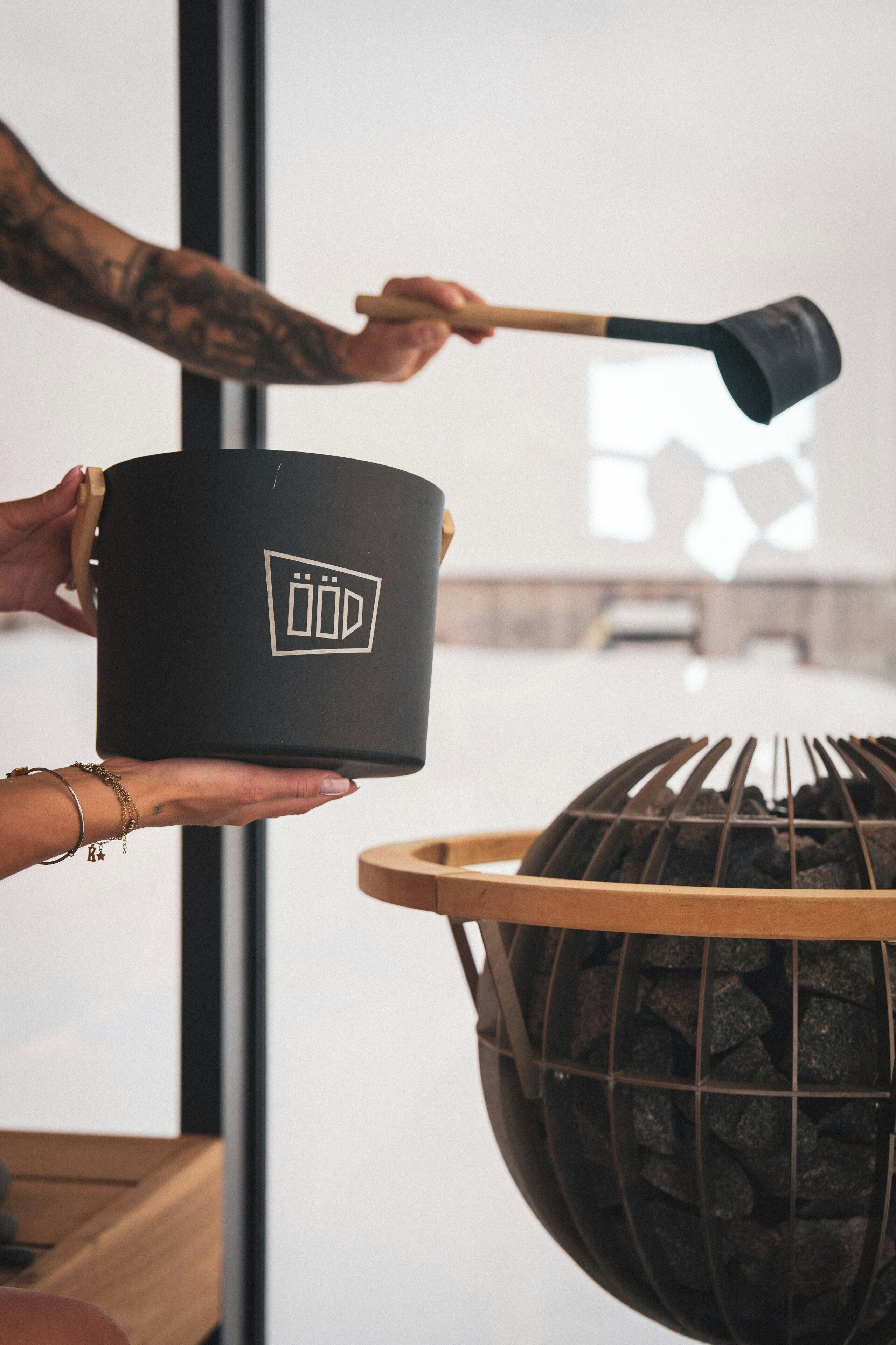 A person holding a black pot with a logo, a grill with charcoal, and a window in the background.