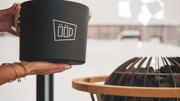 A person holding a black pot with a logo, a grill with charcoal, and a window in the background.