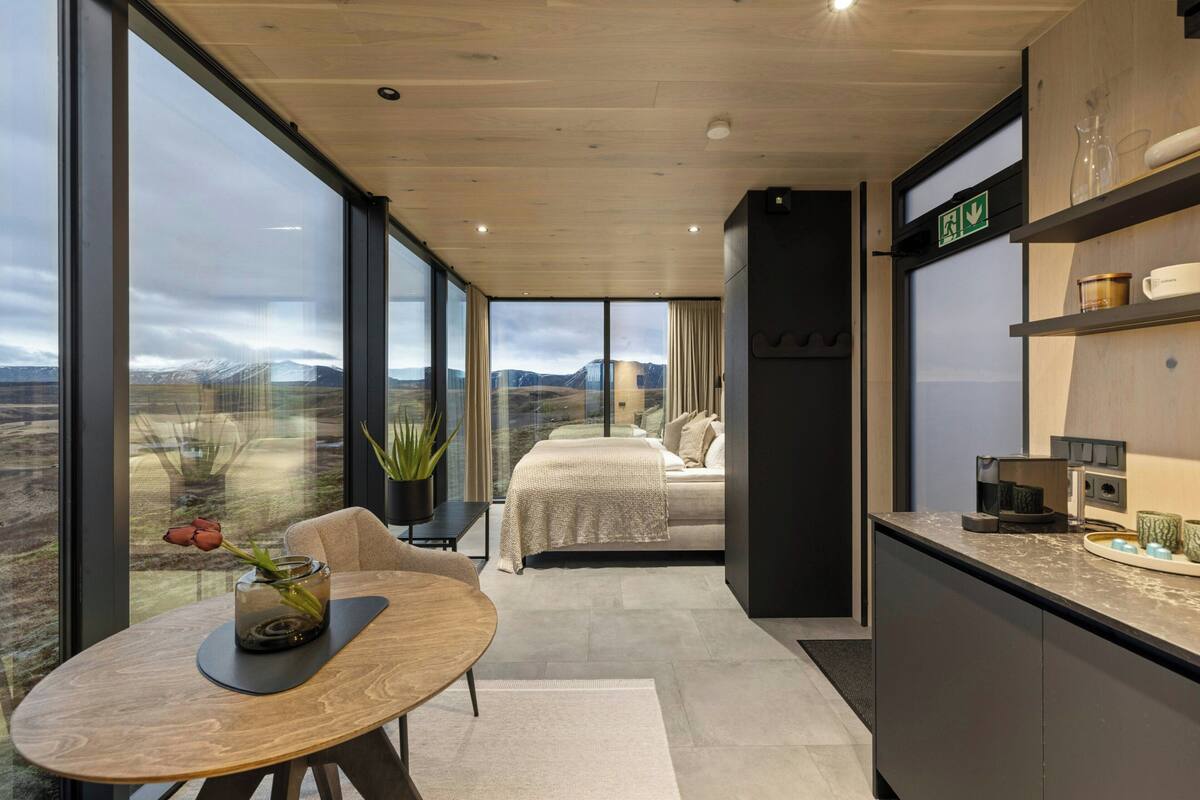 A modern hotel room with a large window, a bed, a dining area, and a kitchen counter.