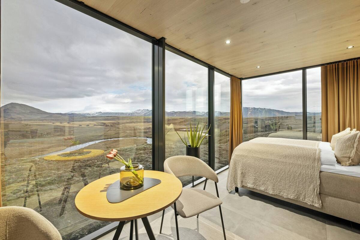 A modern room with a bed, a table with a vase, and large windows offering a mountainous view.
