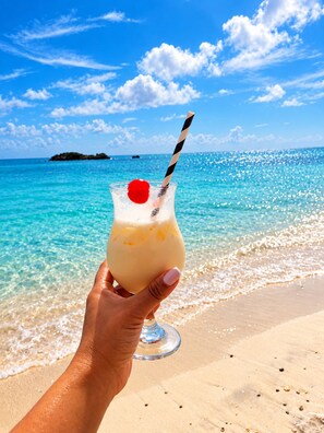 A hand holding a cocktail glass with a cherry on a straw, set against a beach with clear blue water and a distant island. - SHAMAR LODGE  (Providencia)