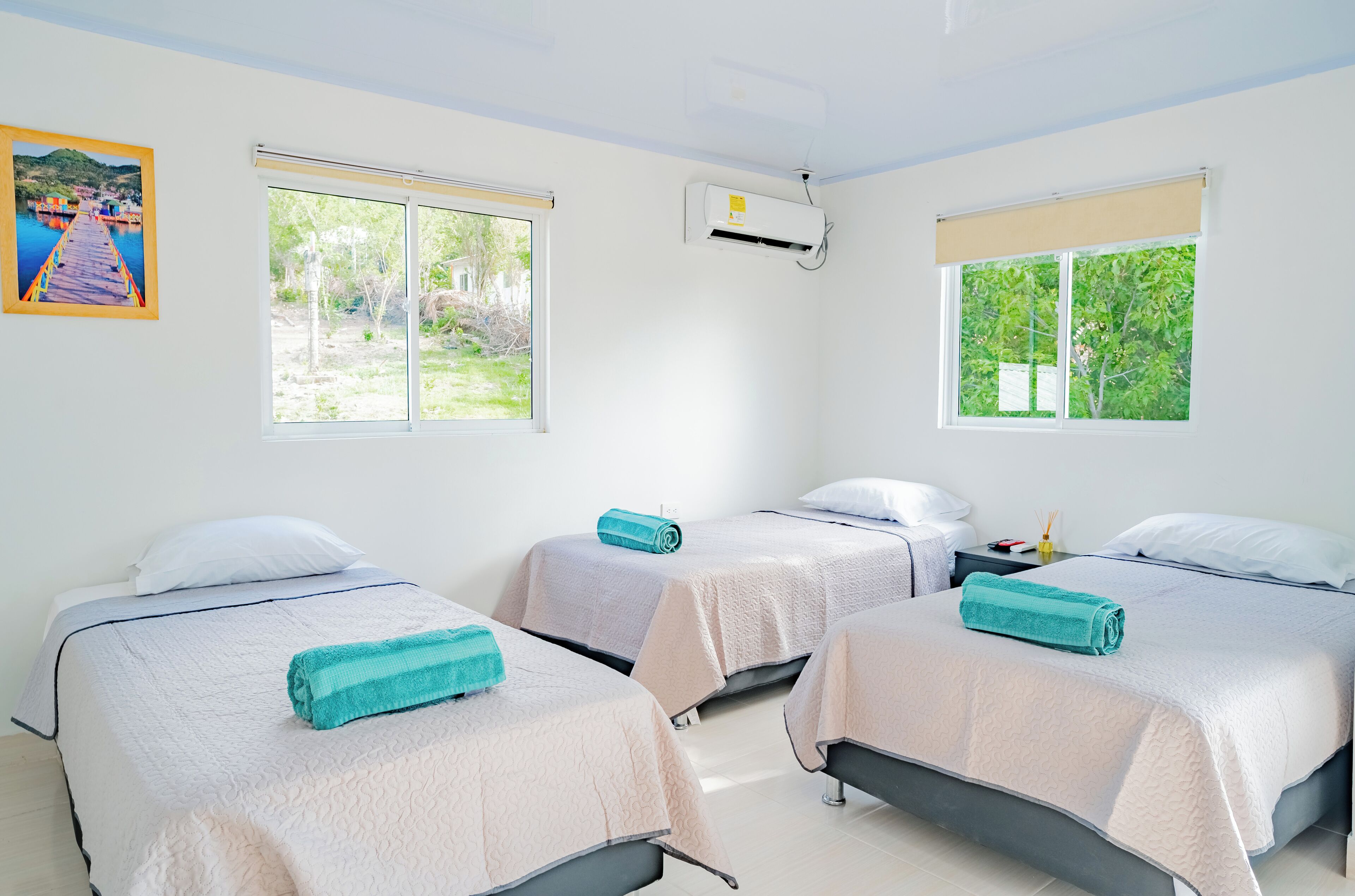 A hotel room with three beds, each with a white bedspread and a teal towel. There are two windows with blinds, one air conditioning unit, and a painting of a pier on the wall.