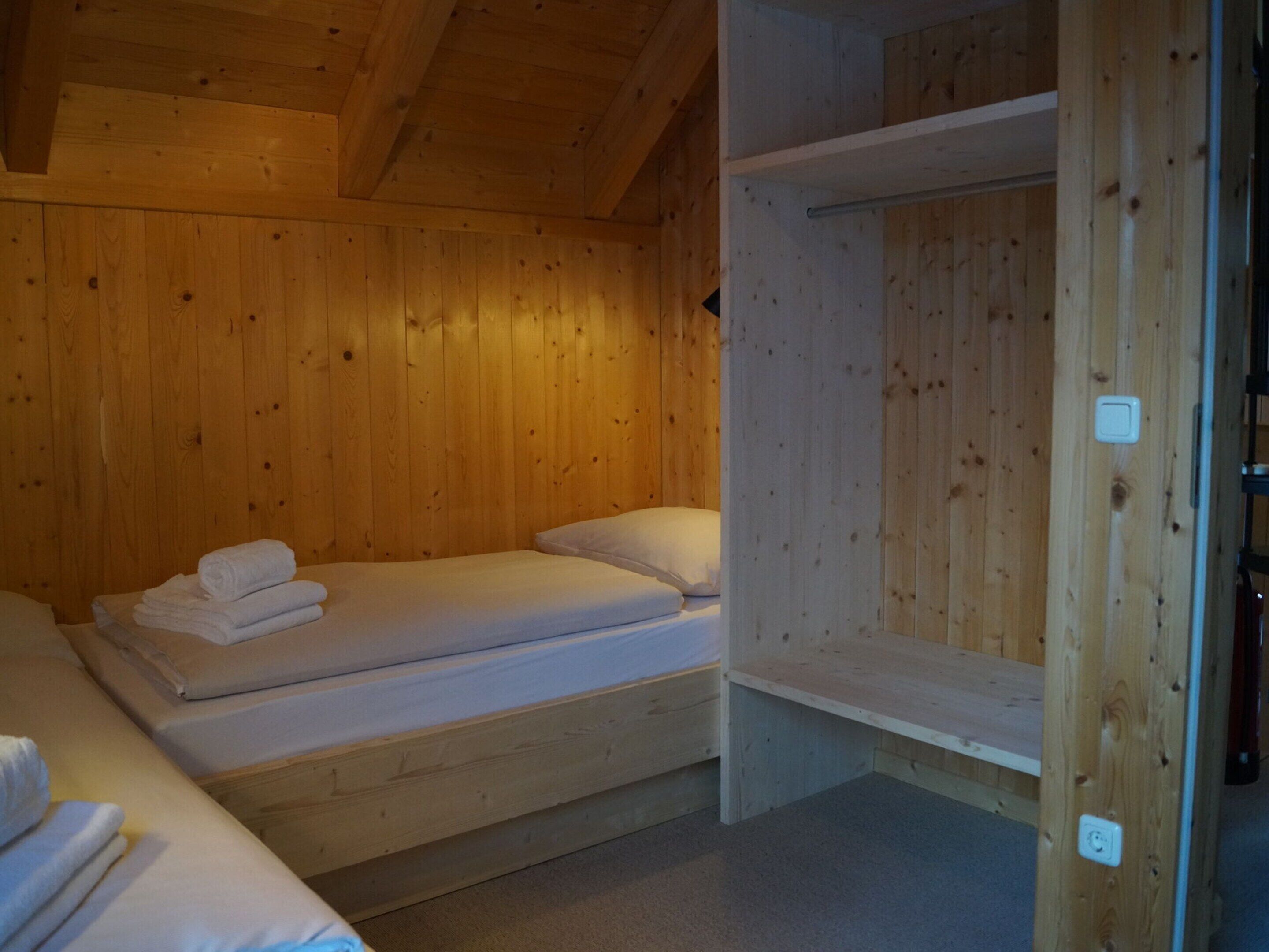 A wooden cabin room with two beds, one with towels on it, and a shelf unit.