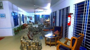 A spacious living area with a patterned sofa, a wooden armchair, and a small table. There are potted plants and a blue trash bin. - Nilanjana Kuakata (Kalapara)