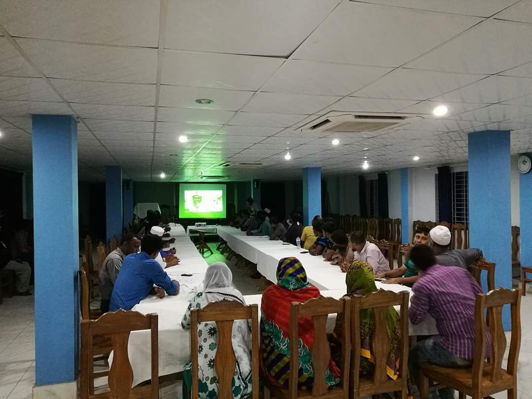 A conference room with a long table and chairs, a projector screen, and people seated facing the screen.